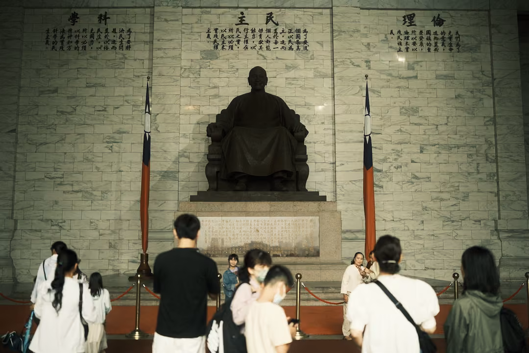 Crowd in white and black clothing behind a red rope, facing a seated bronze statue between two flags