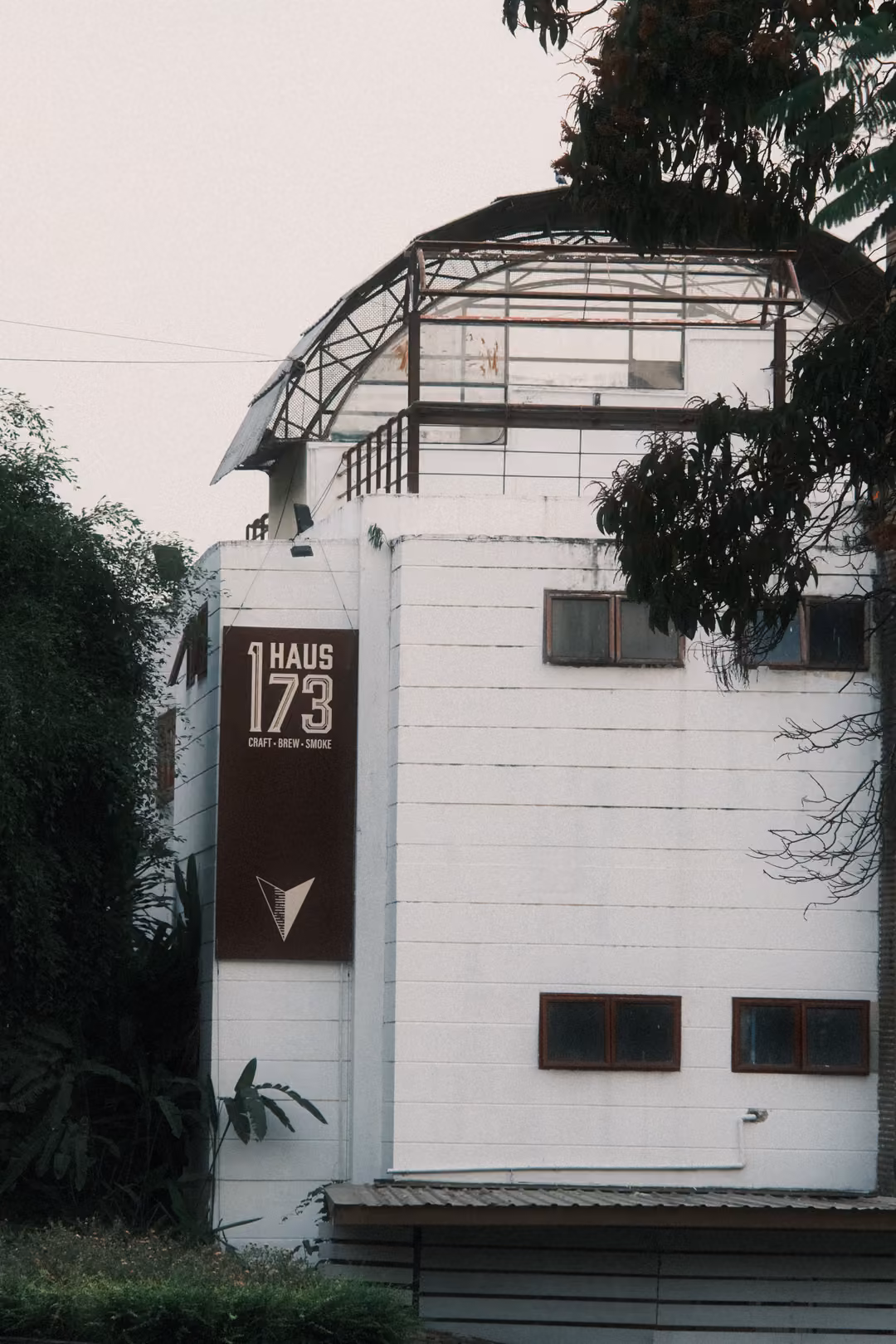 White building with a brown panel reading HAUS 173 and a metal frame on the roof, leaves overhead