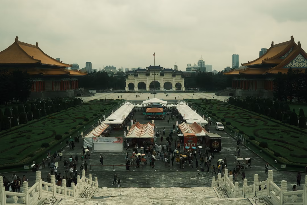 Elevated view over a plaza with white market tents and umbrellas between two orange-roofed halls, grey sky