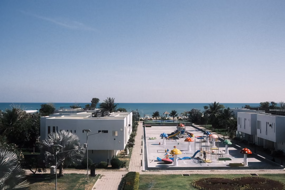 Wide high view of a splash park with colourful umbrellas between white buildings, the sea on the horizon