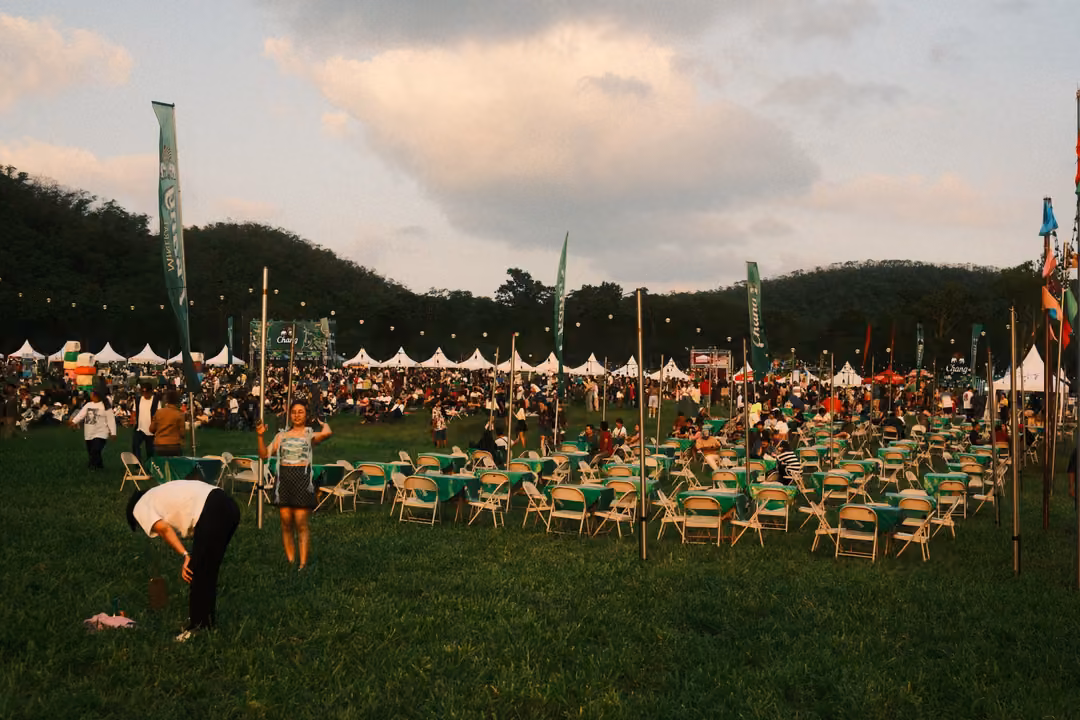 Wide field of green tables and folding chairs below forested hills, with white tents and a few people setting up.
