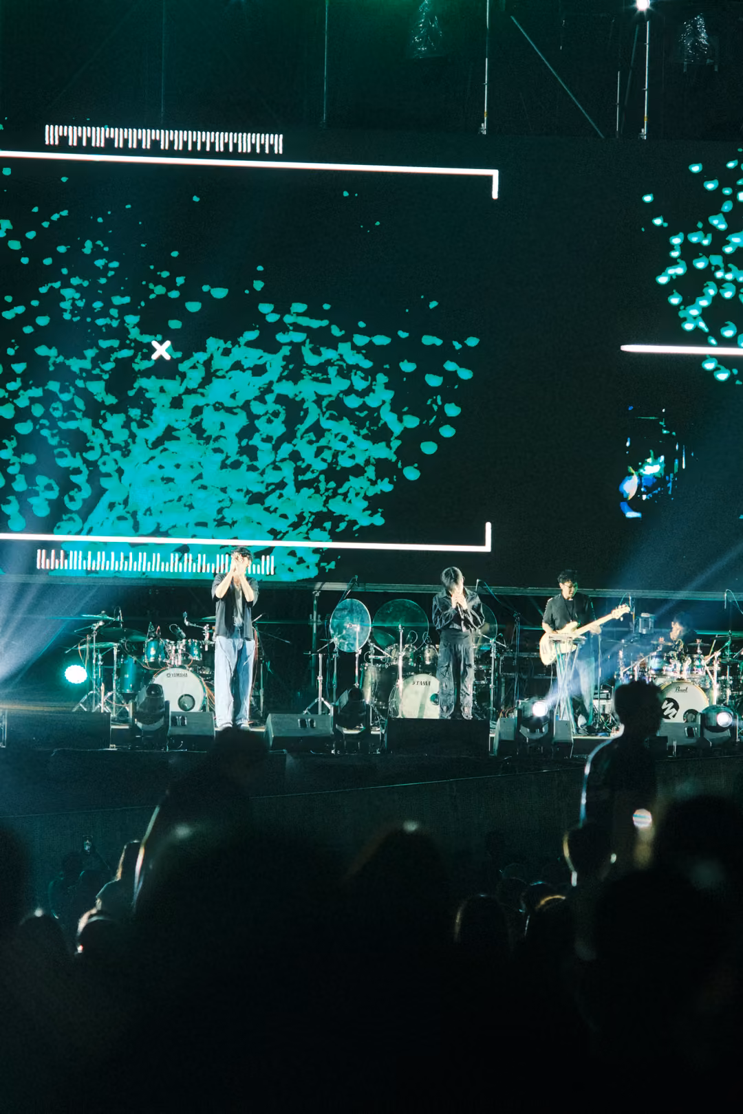 Stage screen covered in a cloud of green dots inside a white bracket, band performing beneath in blue light.