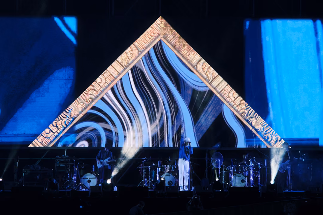 Stage with the singer in a blue jacket under a gold triangle of swirling lines, smoke jets on both sides.