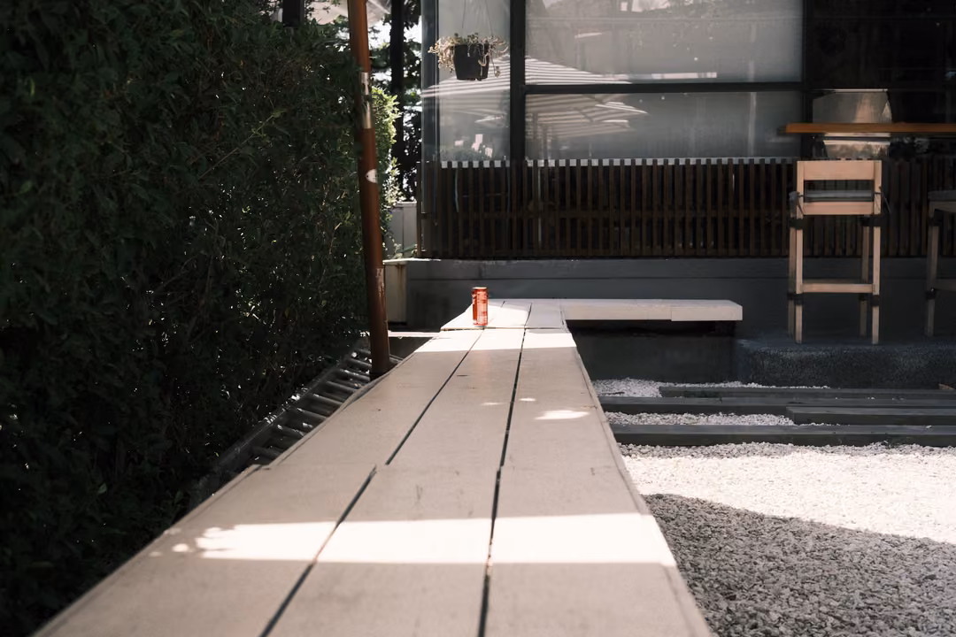 The same wooden boardwalk with a small red can on it, sunlight striping the planks, a glass building behind