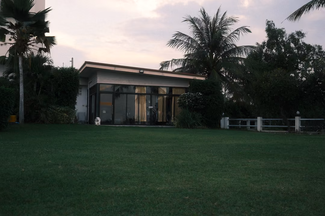 Low modern house with glass windows behind a wide green lawn, palm trees and a pale pink sky