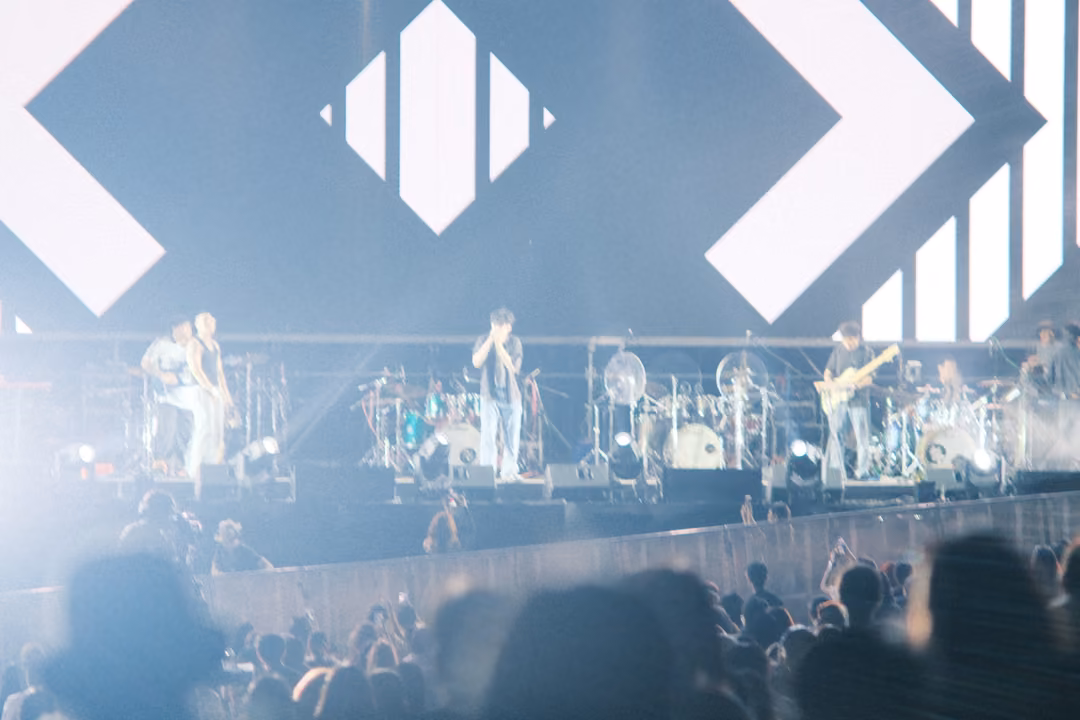 Band on a stage in pale blue haze under giant white chevrons on the screen, crowd silhouettes below.