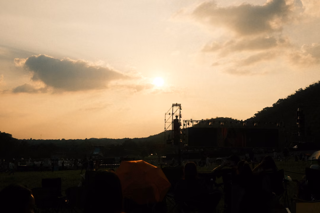 The sun breaks through clouds above a hill, with a silhouetted stage tower and crowd below.