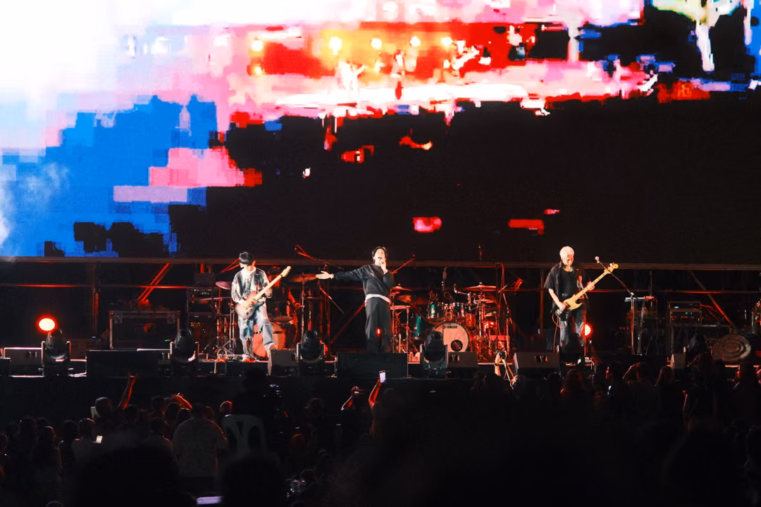 Band on stage below a screen of glitched red, pink and blue pixel blocks, singer gesturing with an arm out.