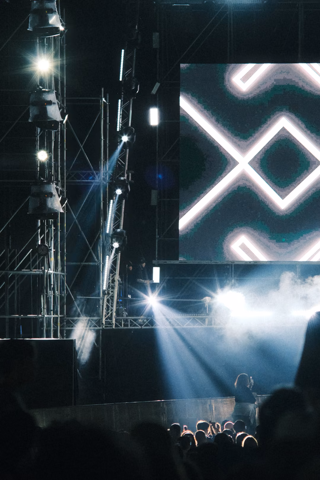 Vertical view of tall scaffolding towers with spotlights beaming into haze, a screen of white X shapes at right.