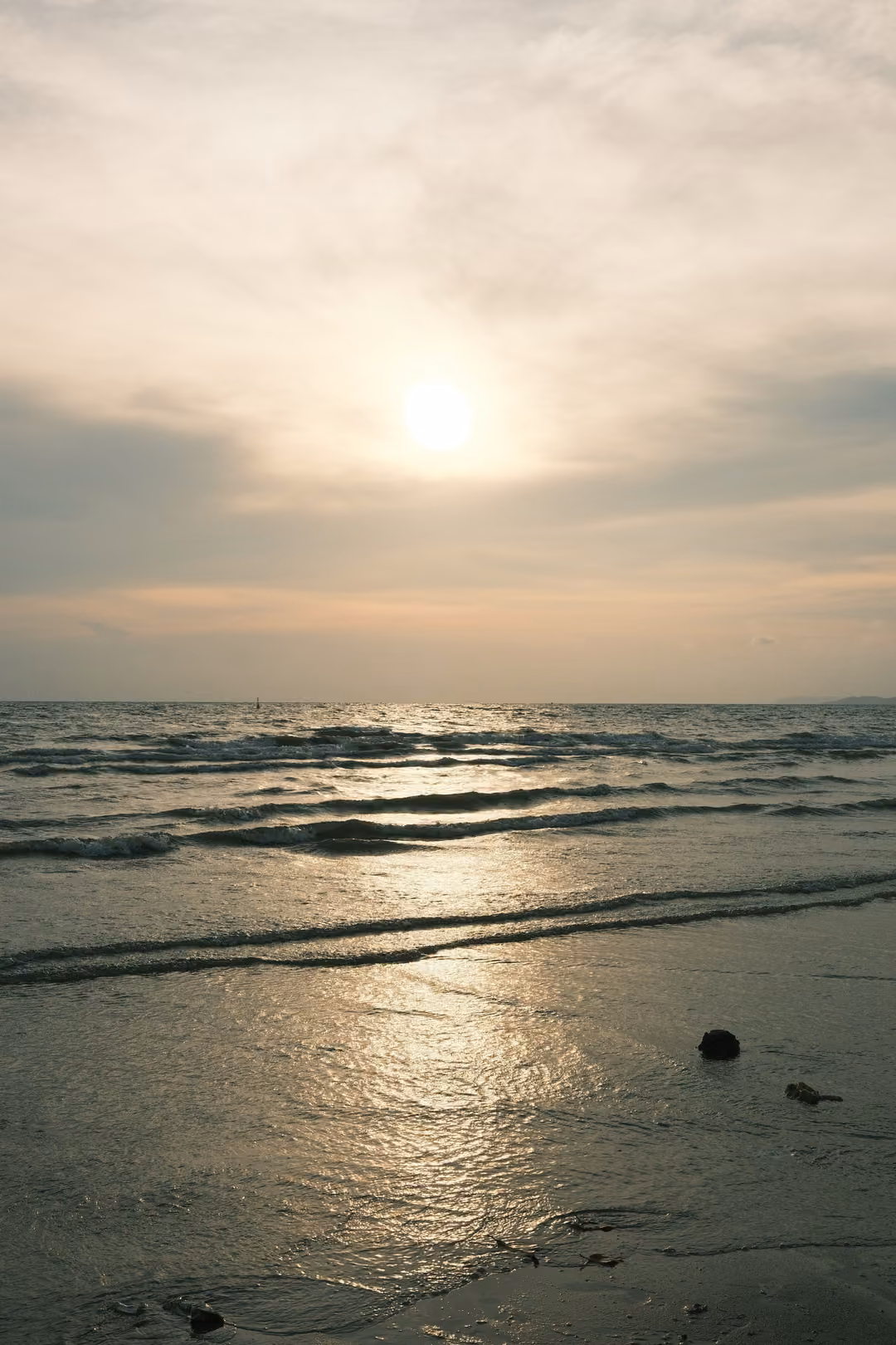 A hazy sun reflected in wet sand at the edge of the sea, a few dark pebbles in the foreground