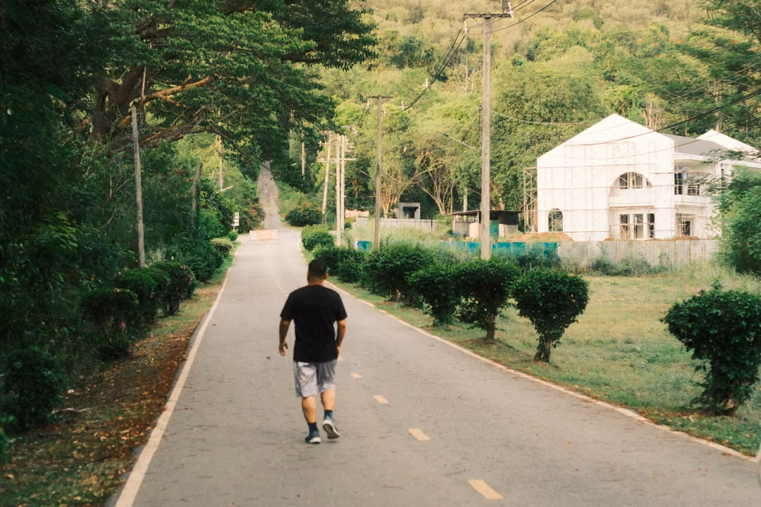 A person in a black T-shirt walks away down a road lined with shrubs toward a white house under construction.