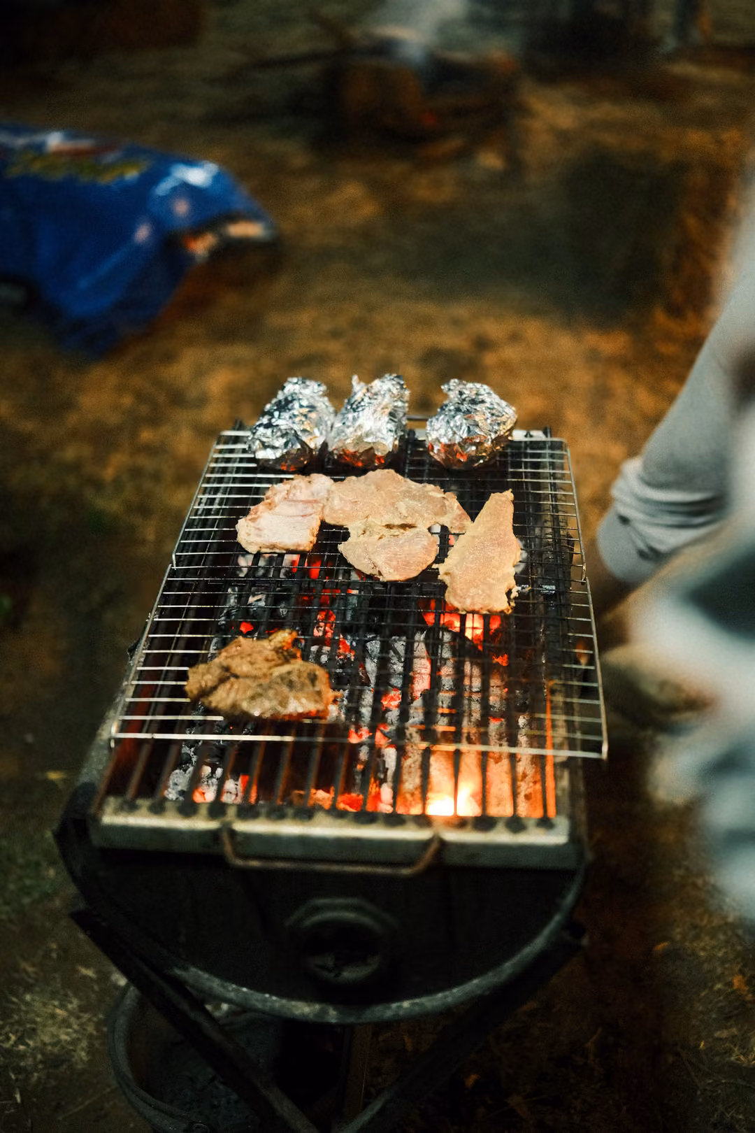 Vertical close view of meat and foil packets on a charcoal grill, coals glowing orange beneath