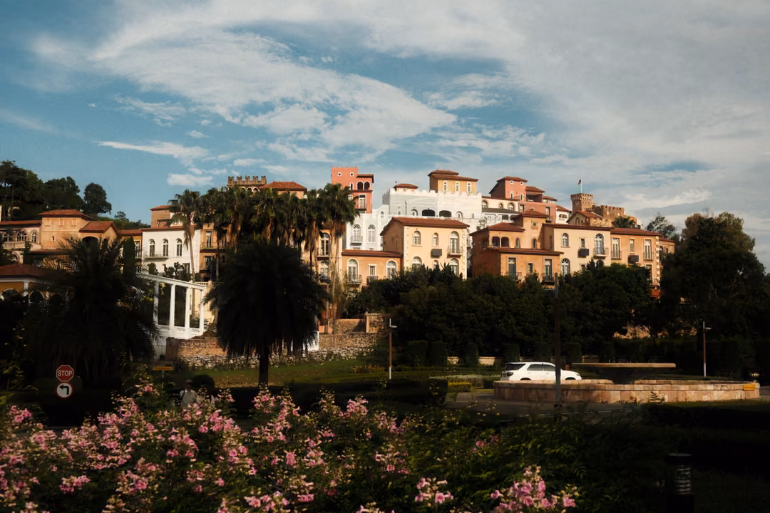 Terracotta and cream buildings on a hillside above palm trees and pink flowers under a blue, cloud-streaked sky