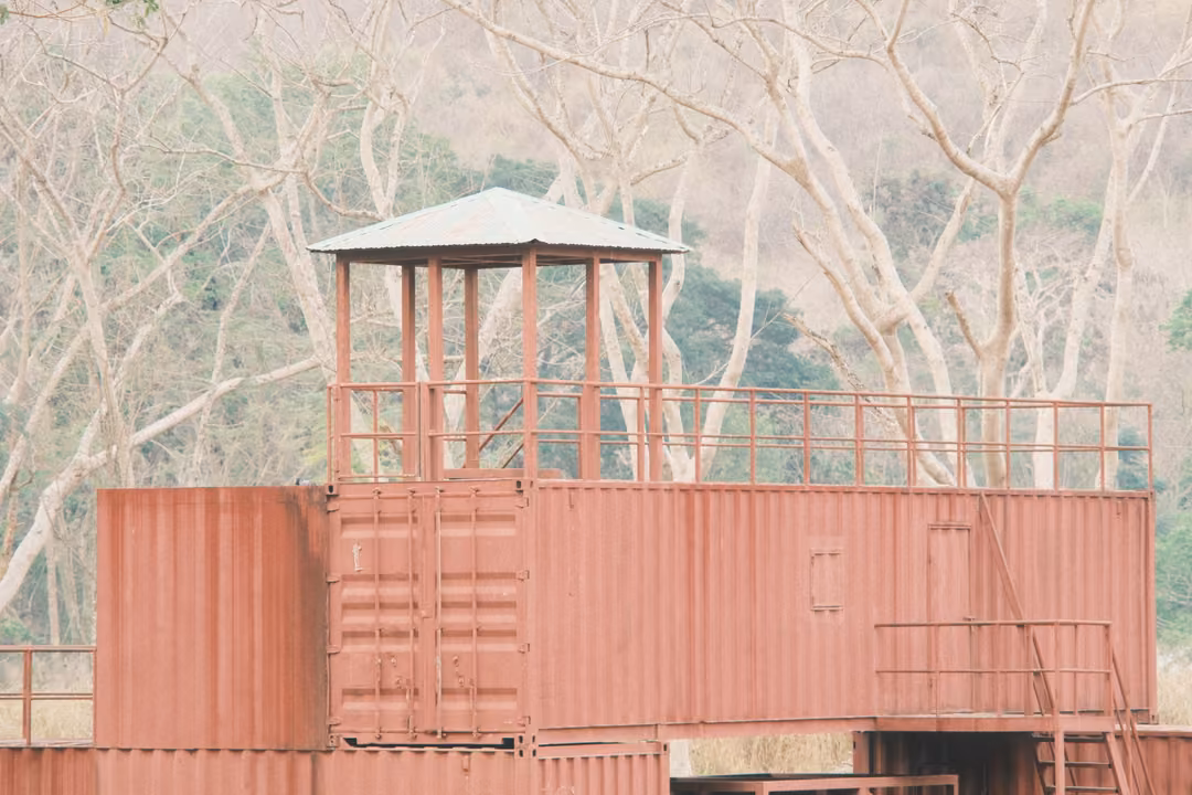 Rust-red structure built from shipping containers with a lookout platform, bare pale trees behind