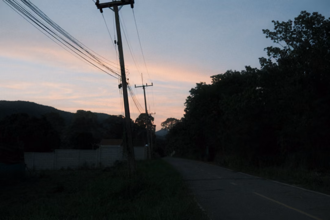 Darker view of a road with power poles against a pink sunset sky, trees and hills in silhouette