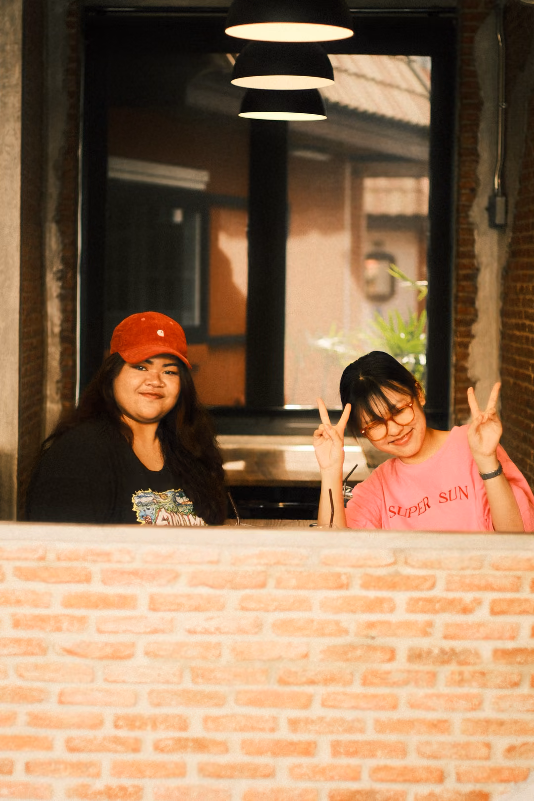 Two women at a cafe window, one in a red cap smiling, one in pink with glasses raising peace signs