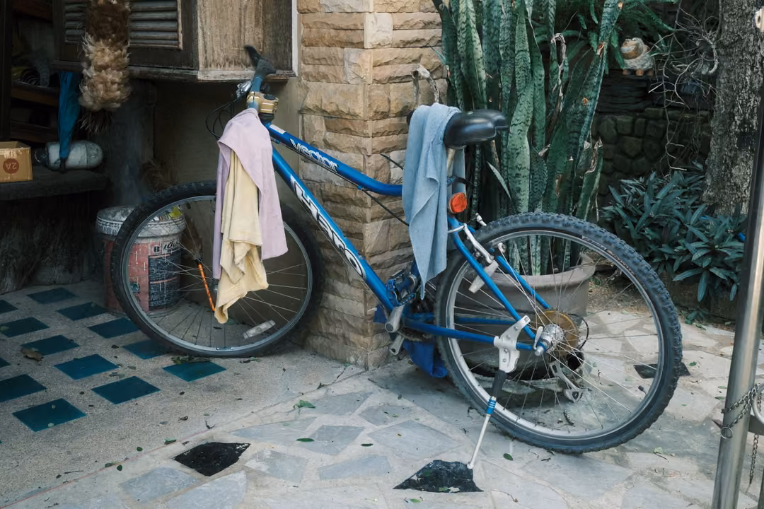 Blue bicycle with towels hung over it parked on a tiled patio by a stone pillar and potted plants