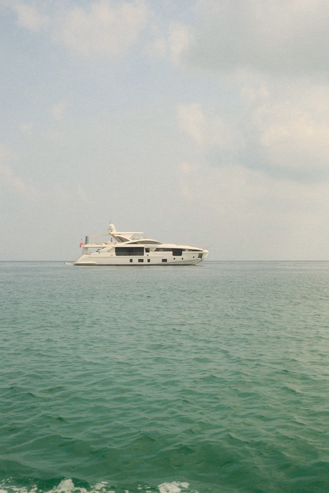 A large white motor yacht anchored on calm green sea beneath a pale sky with scattered clouds.