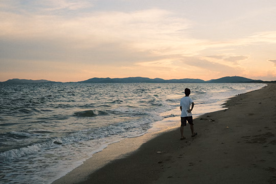 A person in a white T-shirt stands on wet sand facing the sea at sunset, distant hills across the water