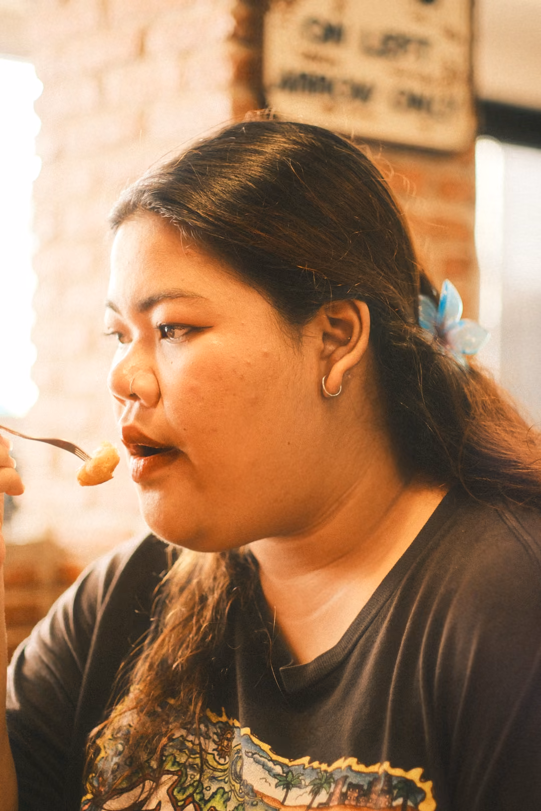 Side profile of a woman with long dark hair eating with a fork, a hair clip and a sign on the brick wall behind