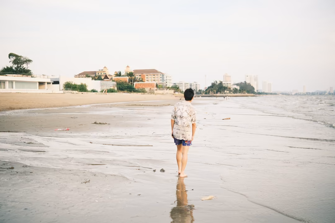 A man in a patterned shirt walking away along the wet beach toward hotels and towers