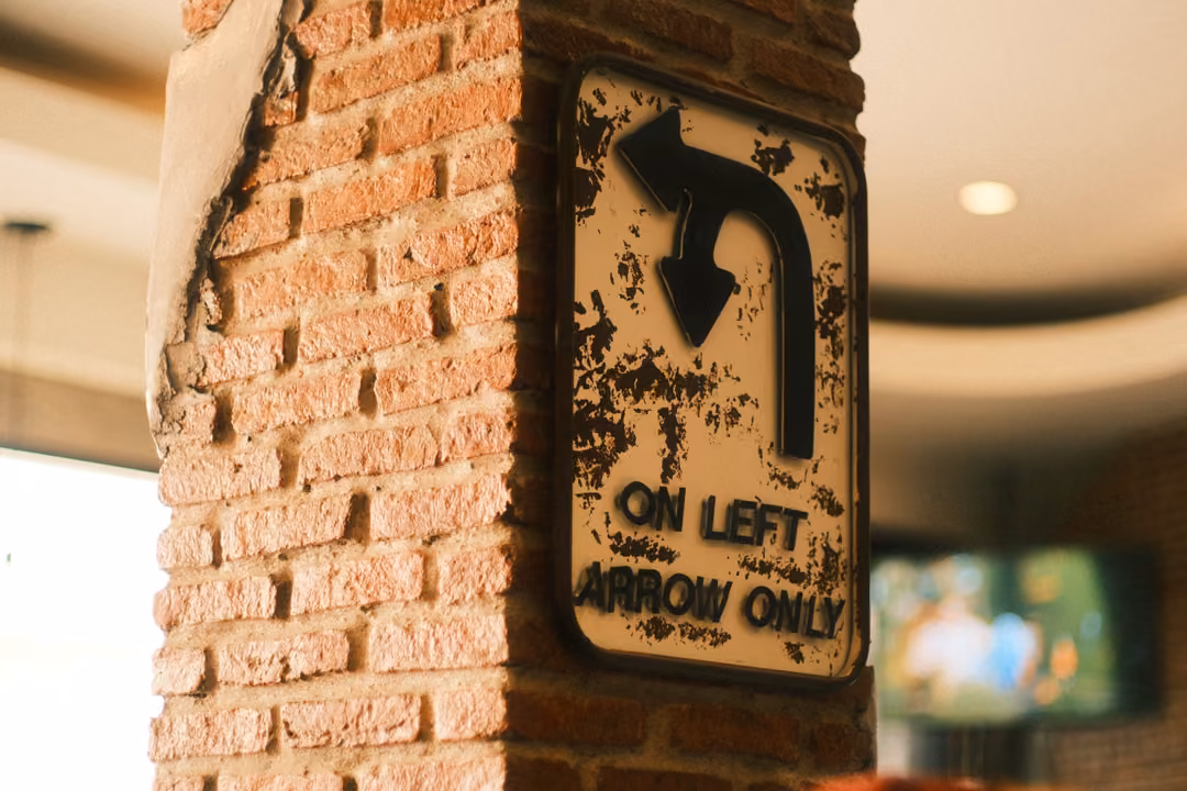 A worn metal road sign on a brick pillar reading ON LEFT ARROW ONLY with a curved turn arrow