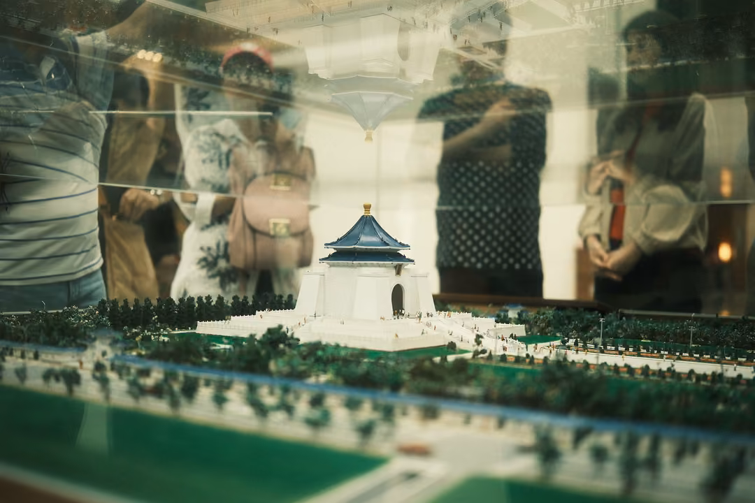 Scale model of a white memorial hall with a blue roof among green lawns, visitor reflections in the glass