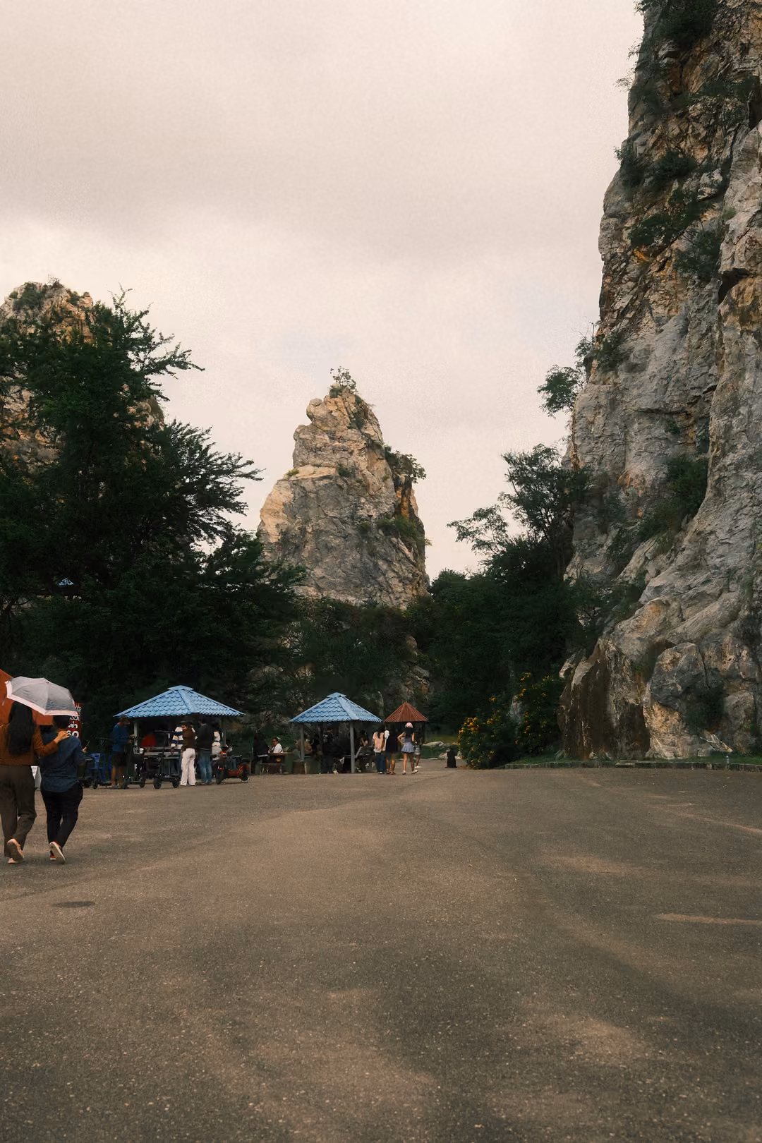 Road leading between steep rock walls toward a spire of limestone, blue umbrellas and small groups of people