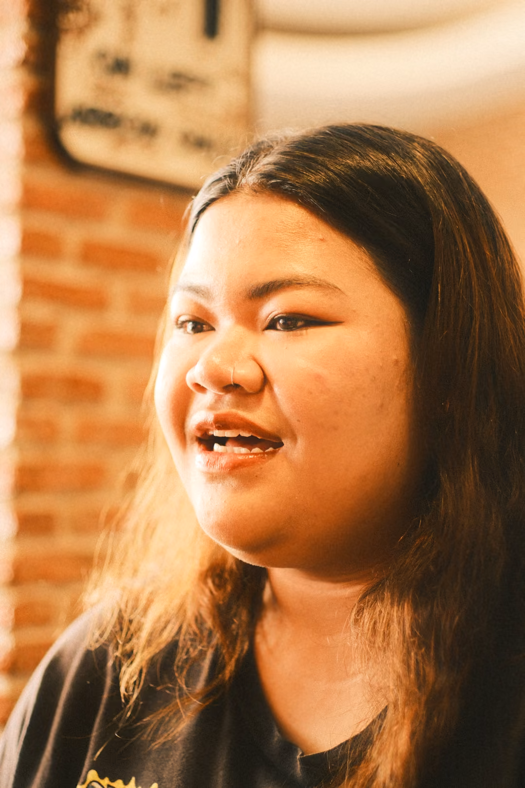 A woman with long hair smiles and talks, warm light on her face, a brick wall and road sign behind