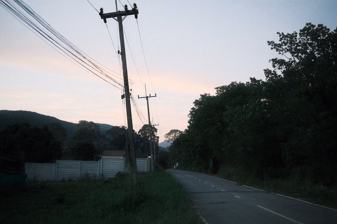 Rural road beside a field with a power pole and wires against a pink evening sky, hills behind